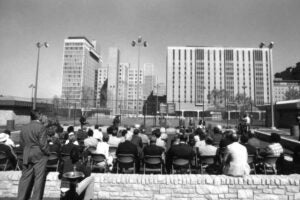 A group of people sit or stand with their backs to us, listening to a speaker in the background standing in front of outdoor tennis courts on a seemingly sunny day.